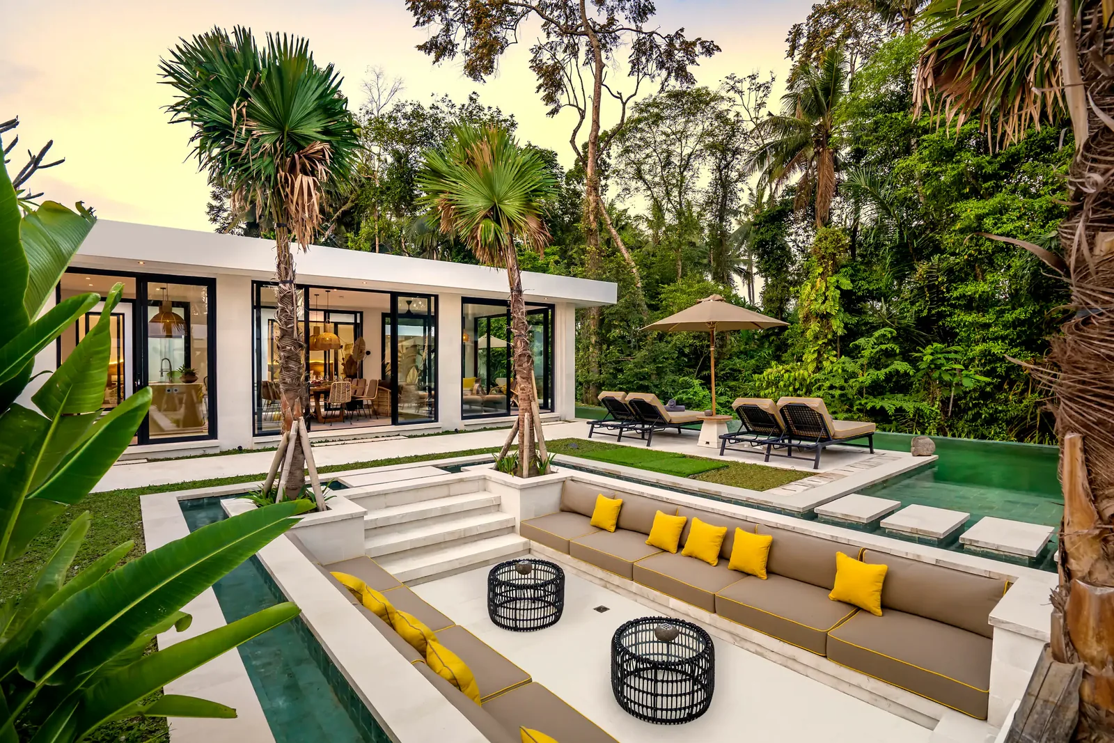 The sunken lounge by the pool, yellow cushions, the house lit behind at dusk